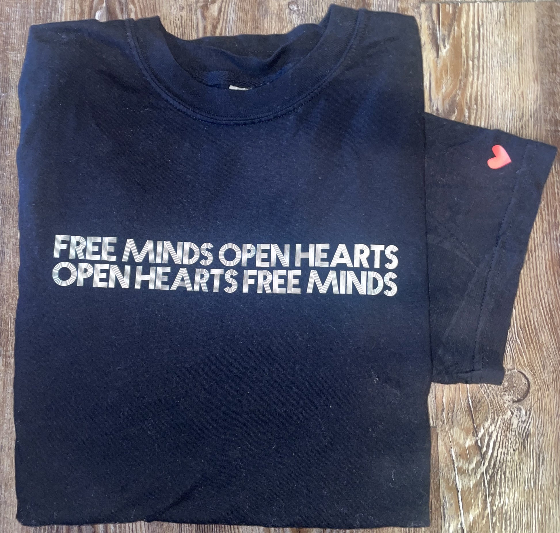 Black Free Minds Open Hearts shirt showing the white text reading "Free Minds Open Hearts Open Hearts Free Minds" printed across the chest, along with a red heart printed on the left sleeve.