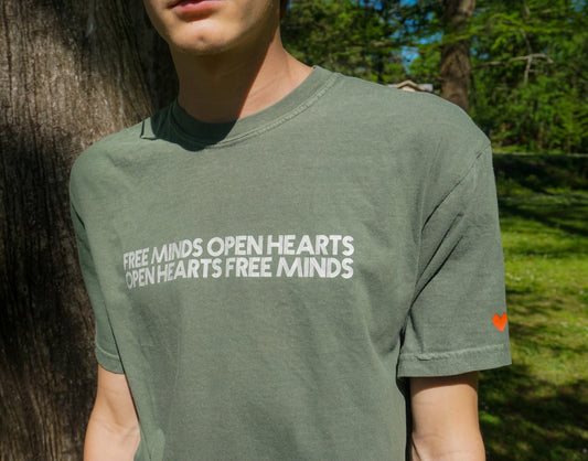 Green Free Minds Open Hearts Shirt shown from the front. Featuring white text reading "Free Minds Open Hearts Open Hearts Free Minds" across the chest, and a red heart printed on the left sleeve.