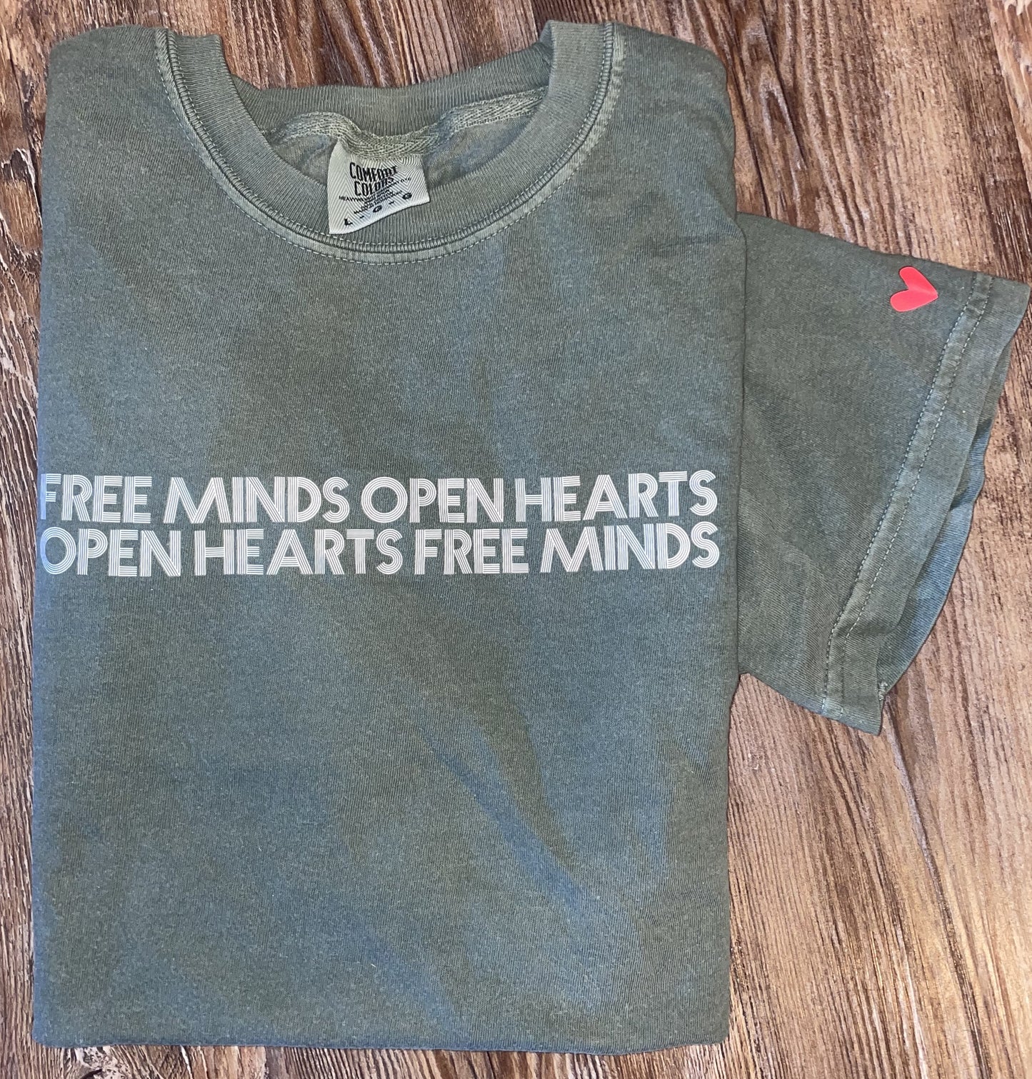 Green Free Minds Open Hearts Shirt shown folded. Featuring white text reading "Free Minds Open Hearts Open Hearts Free Minds" across the chest, and a red heart printed on the left sleeve.